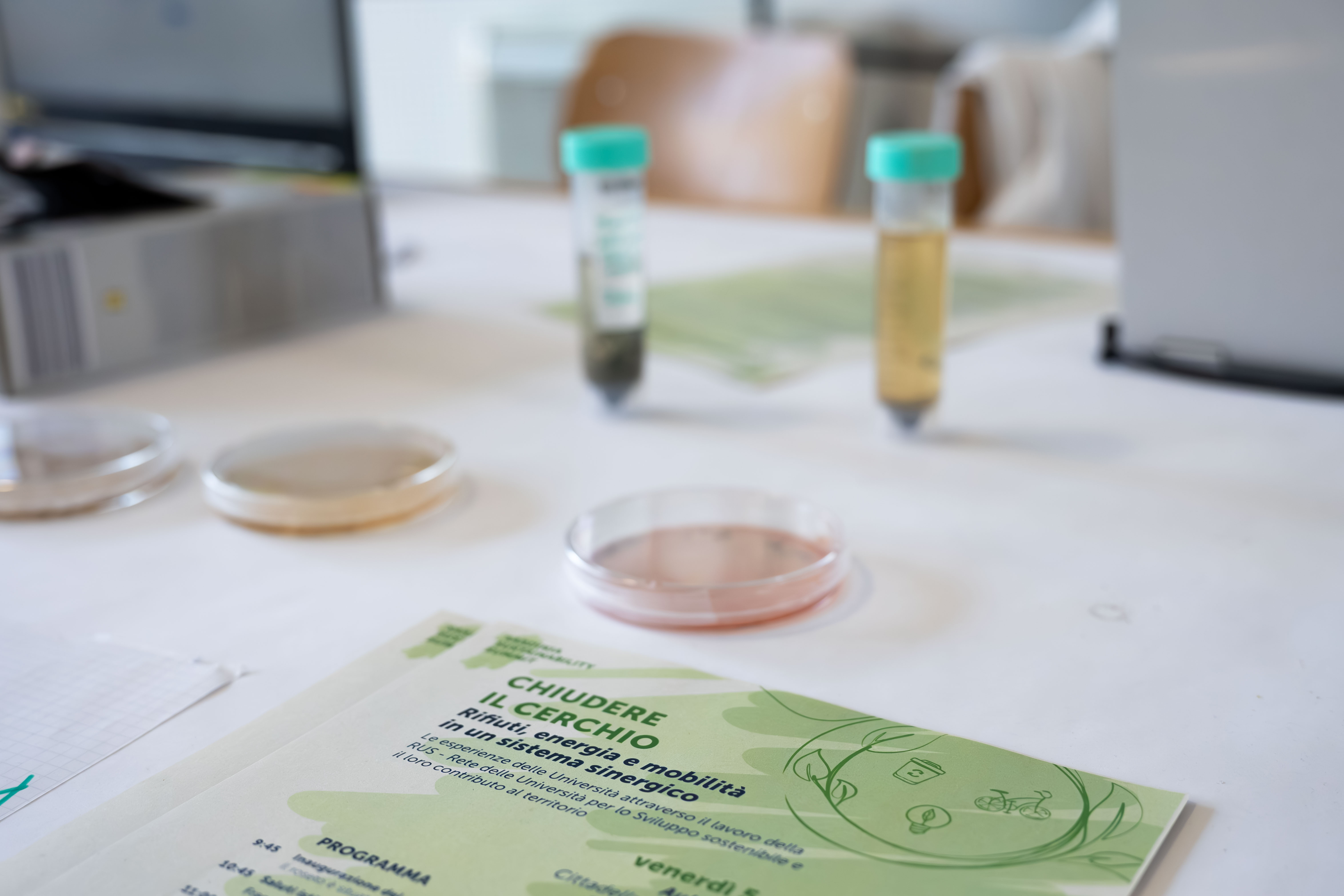 Attività di laboratorio: campioni in provetta e piastre Petri accanto alla locandina dell'evento